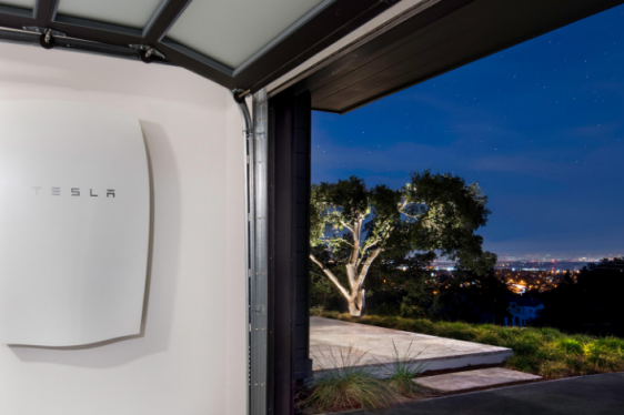 Tesla Powerwal mounted in as a solar battery installation inside a garage with a view of the night sky, city landscape, and trees.